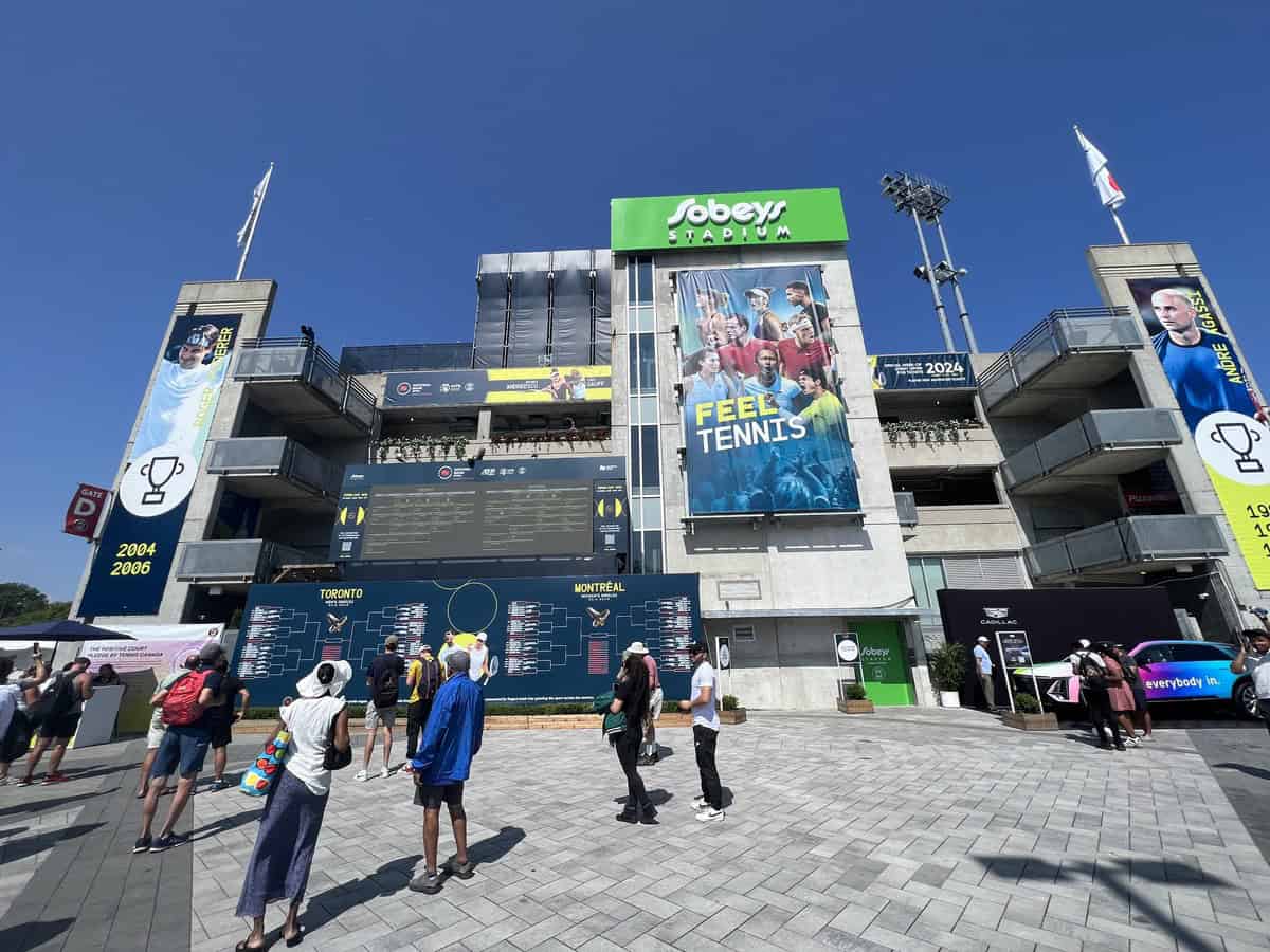 In this image Sobeys Stadium is visible with signage promoting the Canadian Open tennis tournament in Toronto. Fans are reading the large scoreboard visible in front of the stadium. The stadium is three stories in height.