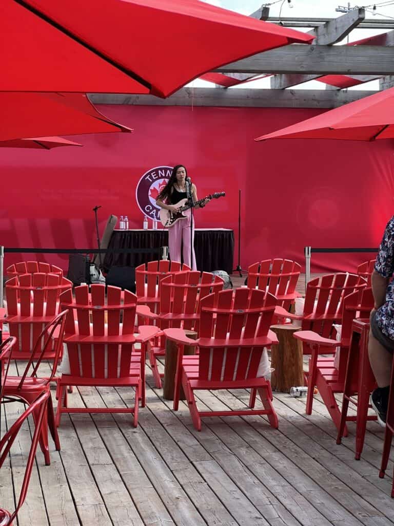 A single performer sings and plays her guitar in an outdoor venue with red muskoka chairs available for fans to sit and enjoy the performance.