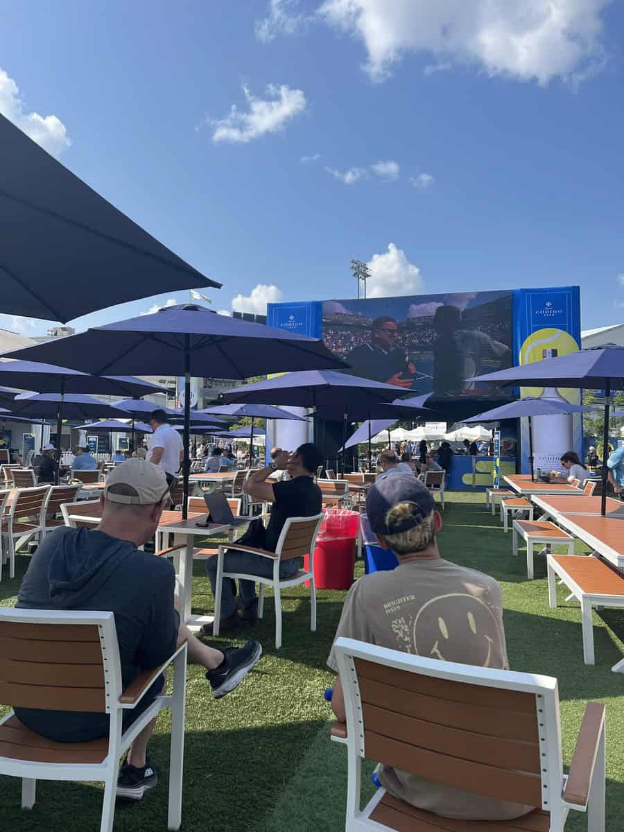 In this image spectators are sitting on an outdoor patio with umbrellas at the National Bank Open Toronto. A large TV screen is visible with the tennis tournament being streamed for fans to watch on the patio.