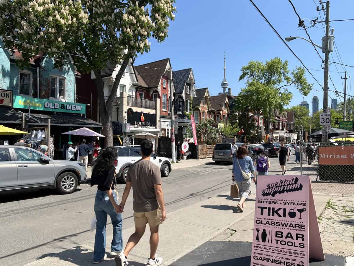 The image shows a lively street in the Kensington Market neighbourhood in Toronto, featuring a mix of shops, people, and vehicles. In the foreground, a couple walks hand-in-hand on the sidewalk. In the background, the CN Tower is visible against a clear blue sky.