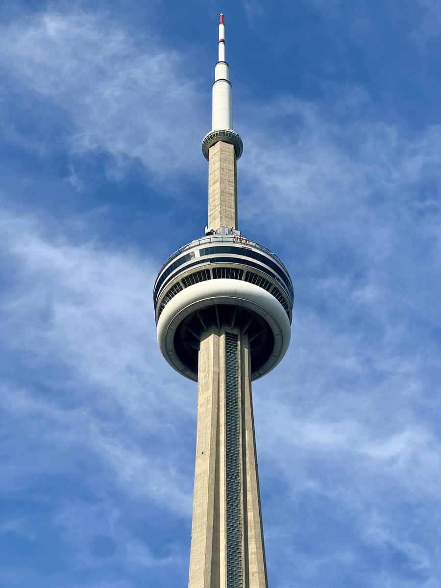 The CN Tower in Toronto, with a bright blue sky as the backdrop. The tower's EdgeWalk attraction is visible, where visitors can be seen walking around the outside ledge near the top.