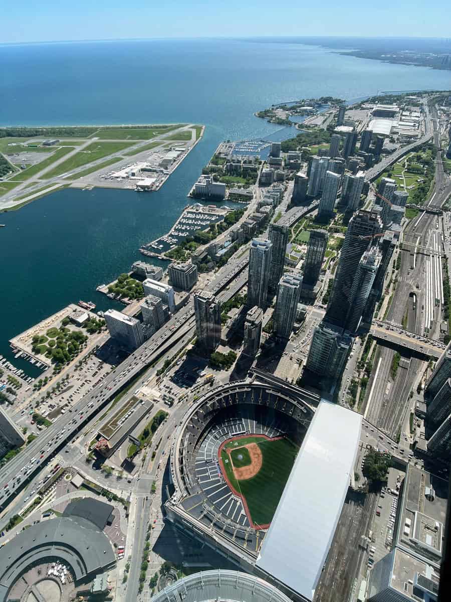 Aerial view from the CN Tower in Toronto, showcasing the Rogers Centre stadium with its retractable roof, the city's waterfront, and the Billy Bishop Toronto City Airport runway. The vast expanse of Lake Ontario stretches out into the horizon under a clear blue sky.