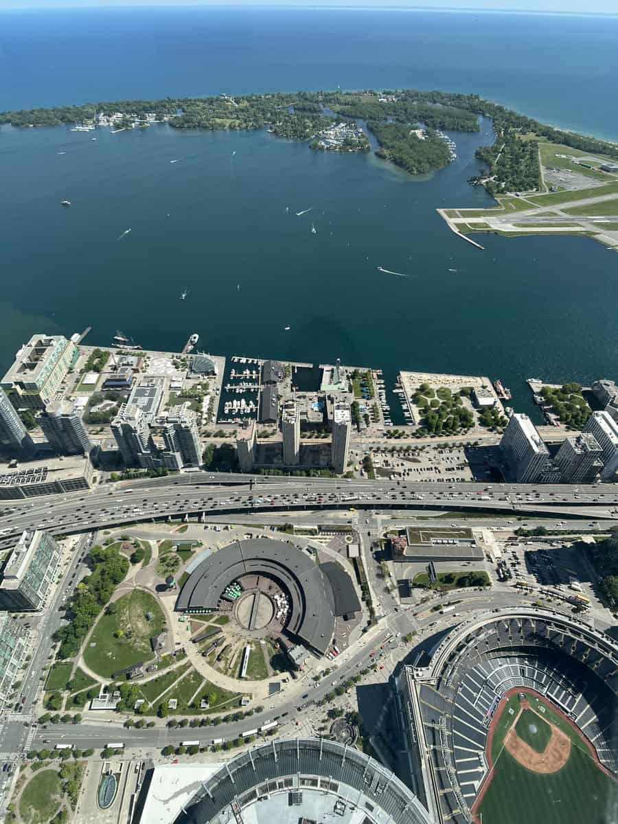 Aerial view from the CN Tower in Toronto, showcasing the Toronto Islands surrounded by Lake Ontario, a busy waterfront with marinas, a round building, and a baseball stadium in the foreground. The green parks and open water create a scenic backdrop against the urban setting.