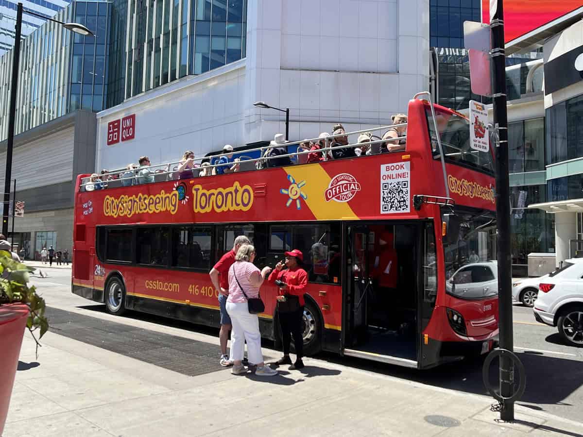 Image of a red City Sightseeing double-decker bus in Toronto, with tourists boarding and seated on the top deck. A staff member in a red uniform assists passengers. The vibrant bus features "Hop On Hop Off" signage and a QR code for online booking, capturing the lively atmosphere of a busy touristic area.