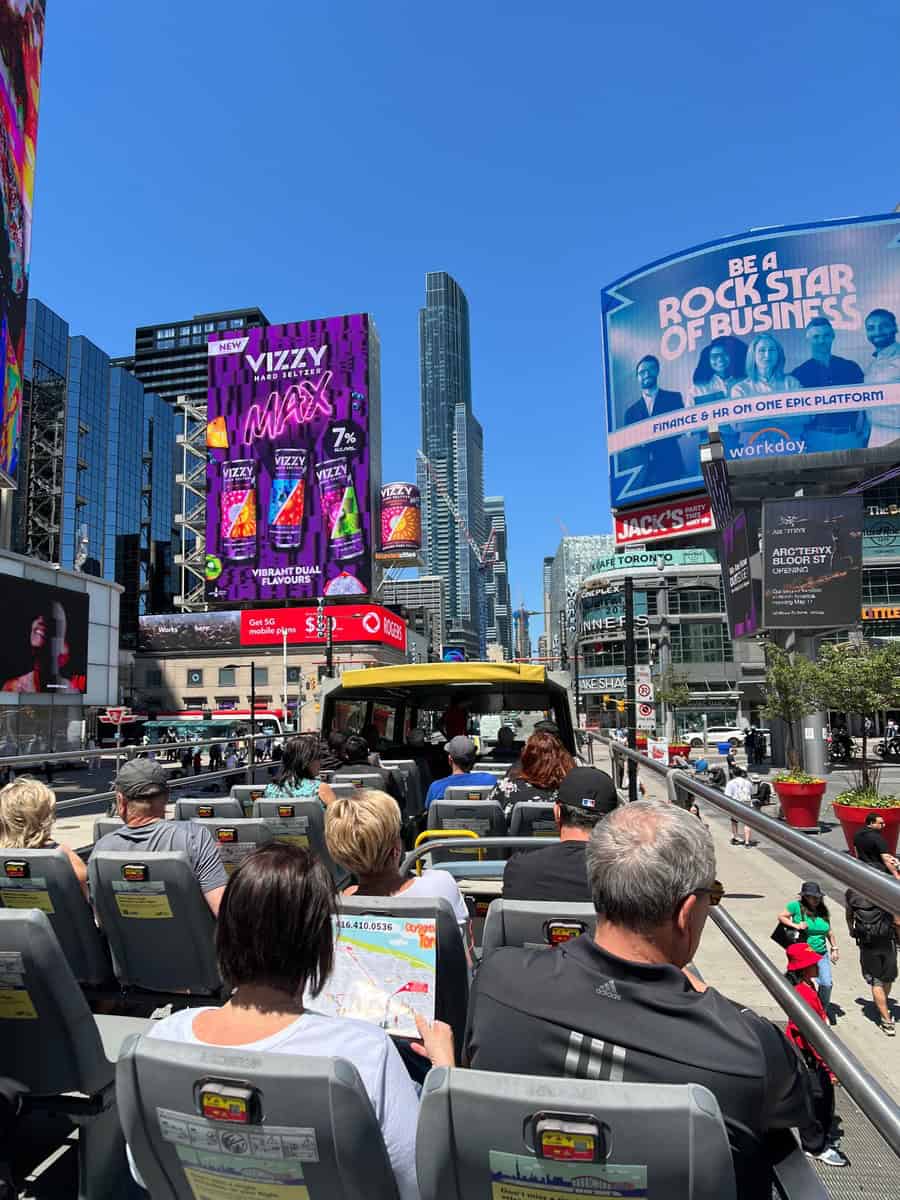 Image from the city sightseeing hop-on hop-off bus in downtown Toronto, with passengers viewing large digital billboards and skyscrapers. The vibrant advertisements dominate the scene against a clear blue sky. The busy urban atmosphere is evident with people walking and traffic in the background.