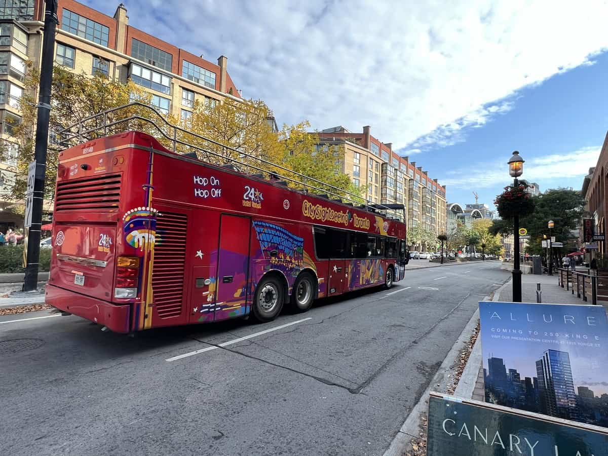 Image of a red City Sightseeing double-decker bus in Toronto, driving through a street lined with modern apartment buildings and trees with autumn foliage. The bus displays "Hop On Hop Off" signage, with colorful graphics, and an advertisement for 24-hour tickets. The sky is partly cloudy, adding a picturesque element to the urban setting.