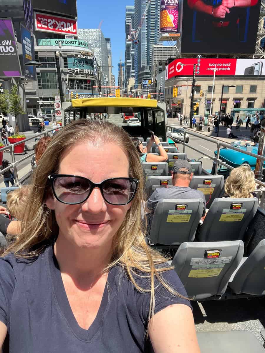 Selfie of a woman wearing sunglasses on an open-top sightseeing bus in downtown Toronto. The bustling cityscape in the background features tall buildings, colorful digital billboards, and a vibrant urban atmosphere under a clear blue sky. The woman is smiling, capturing the excitement of the tour.